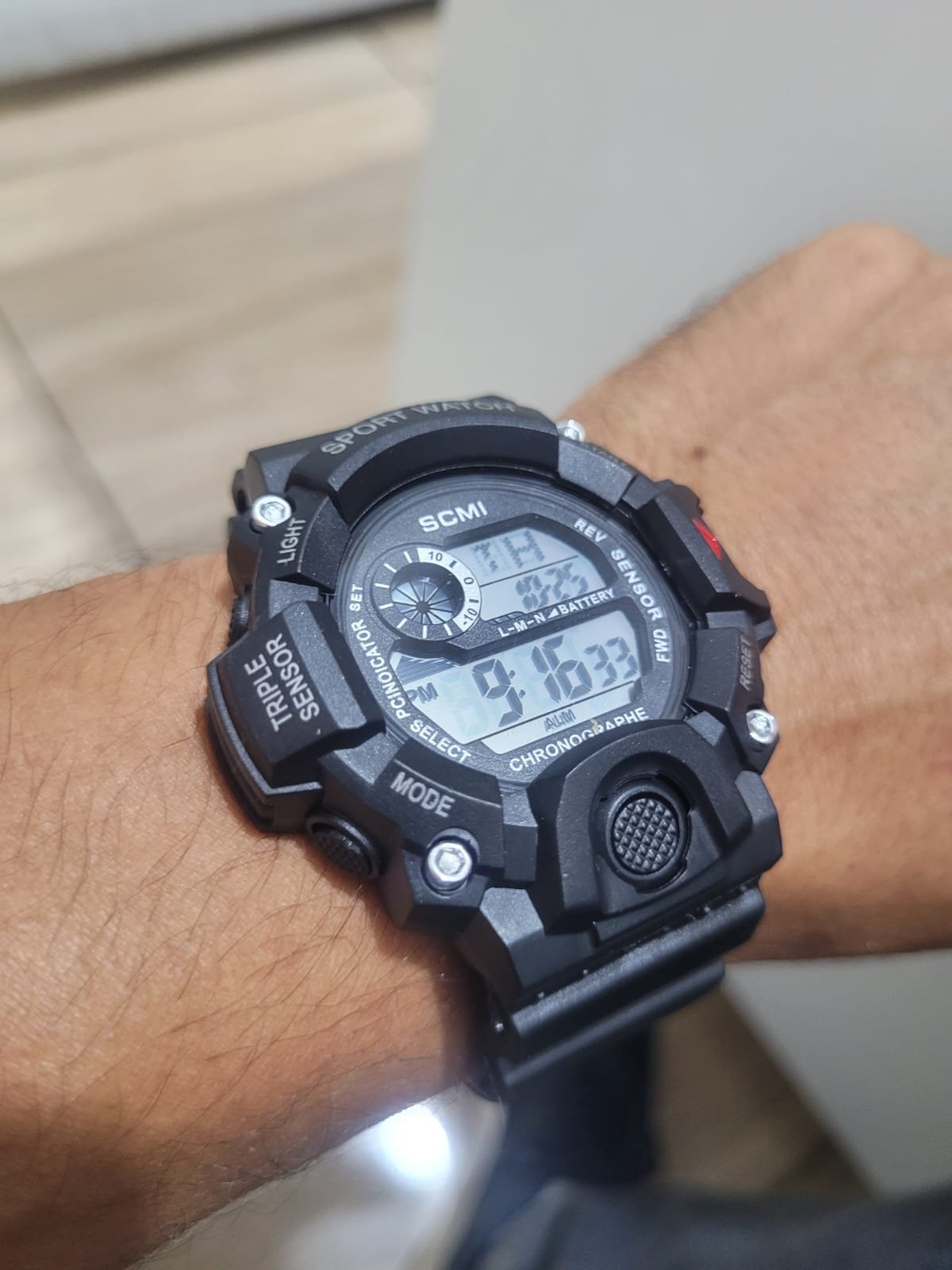 Foto real do relógio digital SCMI preto, com detalhes vermelhos, em sua embalagem transparente